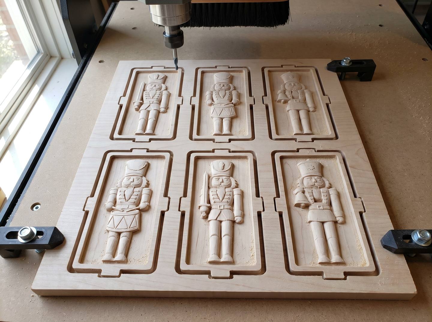 A maple board on a CNC spoilboard with six carved nutcracker ornaments in a neat grid, held by small tabs, with a tapered ball nose bit above