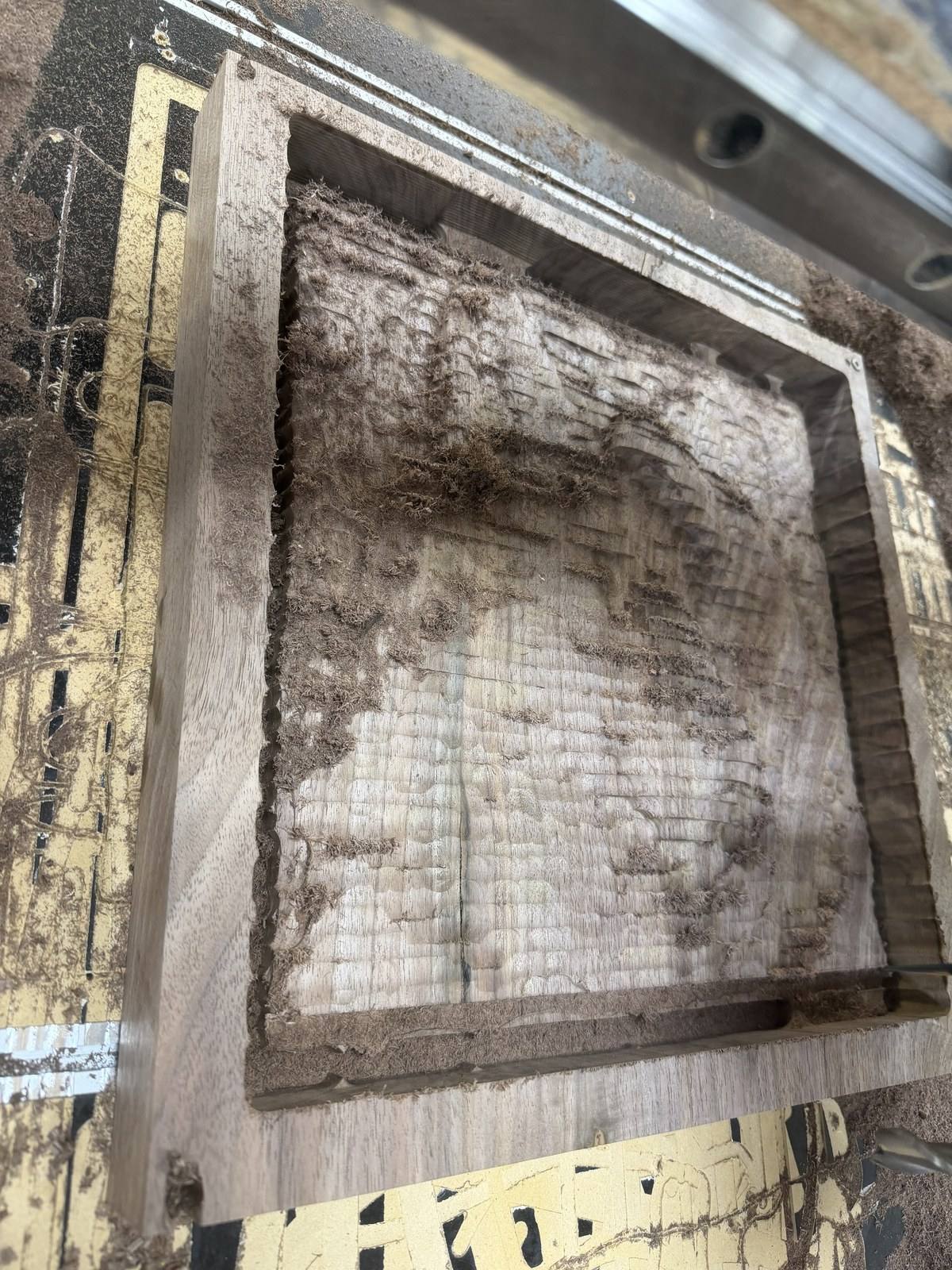 The walnut board after the roughing pass, the wolf and raven shapes cut in broad stepped layers inside a square pocket