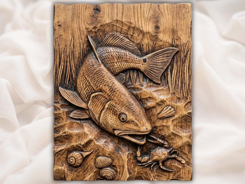 Carved wood relief of a redfish striking toward a blue crab with shells and marsh grass along a rippled shoreline