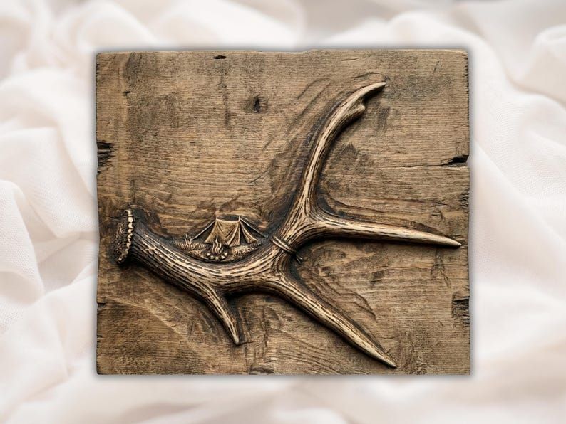 Bas-relief carving of a deer antler with a small tent and campfire scene tucked into the curve, on a rustic wood panel.