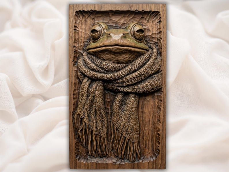 Bas-relief wood carving of a frog with large eyes wrapped in a chunky knitted scarf with fringe.
