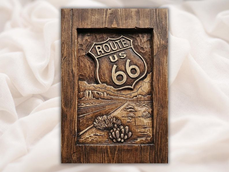 Carved wood relief of the Route 66 highway shield above a desert road with mesas and a cactus.