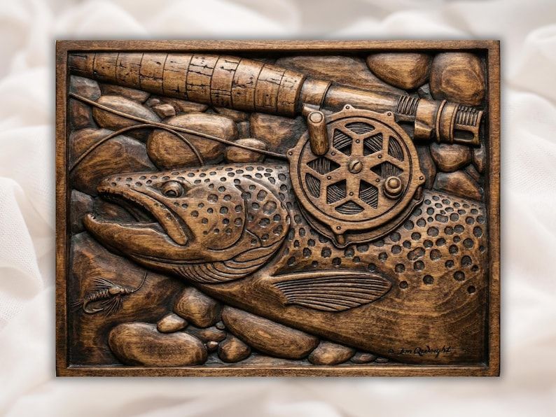 Carved wood relief of a brown trout with a fly fishing rod, reel and mayfly resting on river stones